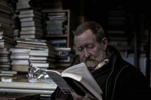 Elderly man reading a book with piles of books in the background